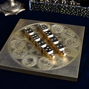 May include: A gold-colored game board with a circular design of embossed coins. Two gold dice are placed on the board. The board is on a dark blue surface. Other gold objects are in the background.