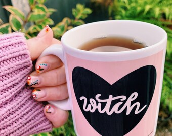 May include: A white ceramic mug with a pink background and a black heart design. The word "witch" is written in white script inside the heart. The mug is filled with tea and held by a person with decorated nails.