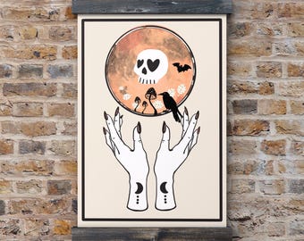 May include: A framed art print featuring a skull with heart-shaped eyes, a bat, and a raven. The skull is set against an orange and brown background. White hands with long black nails and crescent moon designs are at the bottom.