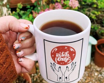 May include: White ceramic mug filled with dark liquid, possibly coffee or tea. The mug features a graphic with the words "witch please" in a red circle, with hands reaching up. The mug is held by a person with decorated nails.