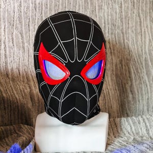 May include: Black balaclava with a white spiderweb design. The eye openings are outlined in red and have a blue mesh. The balaclava is displayed on a white mannequin head.