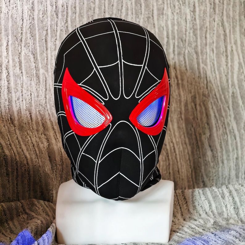 May include: Black balaclava with a white spiderweb design. The eye openings are outlined in red and have a blue mesh. The balaclava is displayed on a white mannequin head.
