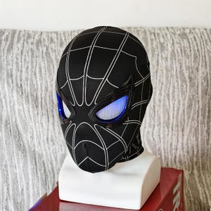 May include: Black Spider-Man mask with white web detailing. The mask features blue eye lenses and is displayed on a white mannequin head. The mask is made of a flexible material.