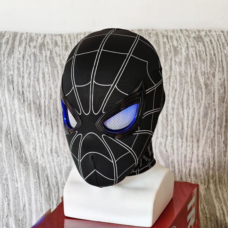 May include: Black Spider-Man mask with white web detailing. The mask features blue eye lenses and is displayed on a white mannequin head. The mask is made of a flexible material.