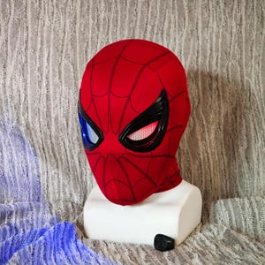 May include: A red Spider-Man mask with black web detailing and black eye frames. The eyes have blue and white lenses on the left and red and white lenses on the right. The mask is displayed on a white mannequin head.