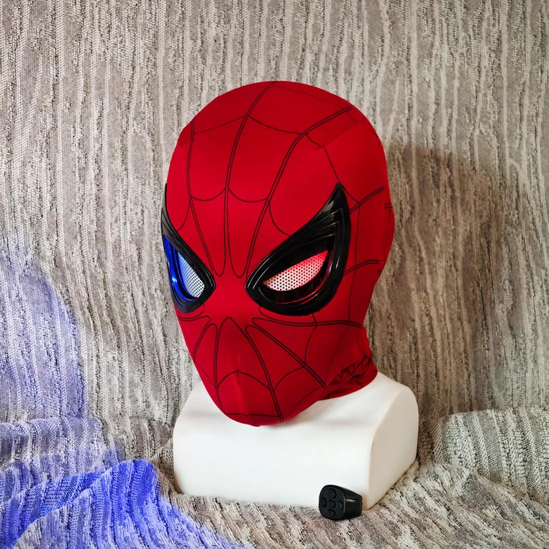 May include: A red Spider-Man mask with black web detailing and black eye frames. The eyes have blue and white lenses on the left and red and white lenses on the right. The mask is displayed on a white mannequin head.