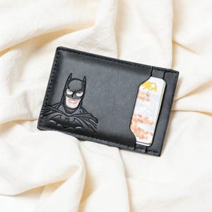 May include: Black cardholder featuring a raised Batman graphic. The wallet has a slot with a card partially visible. The cardholder is made of a smooth material and is set against a white fabric background.