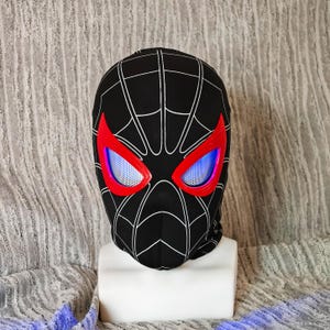 May include: Black balaclava with a white web pattern. The eye openings are outlined in red with blue mesh inserts. The balaclava is displayed on a white mannequin head.