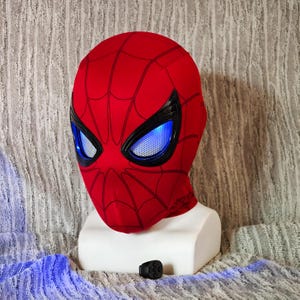 May include: A red Spider-Man mask with black webbing and eye frames. The eyes have a blue, glowing effect. A small black remote control is visible below the mask. The mask is displayed on a white bust.