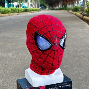 May include: A red Spider-Man mask with black web detailing and large, reflective eye lenses. The mask is displayed on a white stand, with the base of the stand resting on a box with the text "THE AMAZING SPIDER-MAN 2".