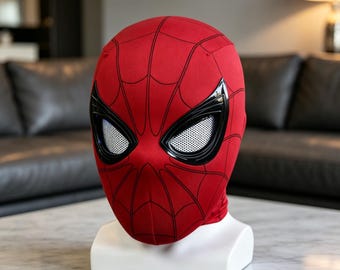May include: A red fabric mask featuring a black web pattern across the surface. The mask includes two large, angular eye lenses with black frames and white mesh inserts, displayed on a white stand to showcase the full head shape and design.