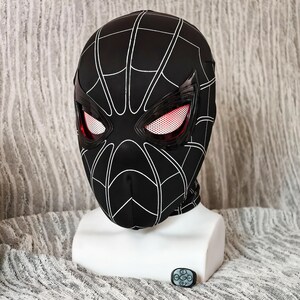 May include: A black Spider-Man mask with white web detailing. The mask has red and white eye lenses and is displayed on a white mannequin head. A small black remote control is in front of the mannequin.