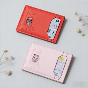 May include: Two leather cardholders, one red and one pink, each featuring a cat-themed design. The red cardholder has a cutout of a cat mask and the text "CAT ID". The pink cardholder has a similar design. Both have a slot for cards.
