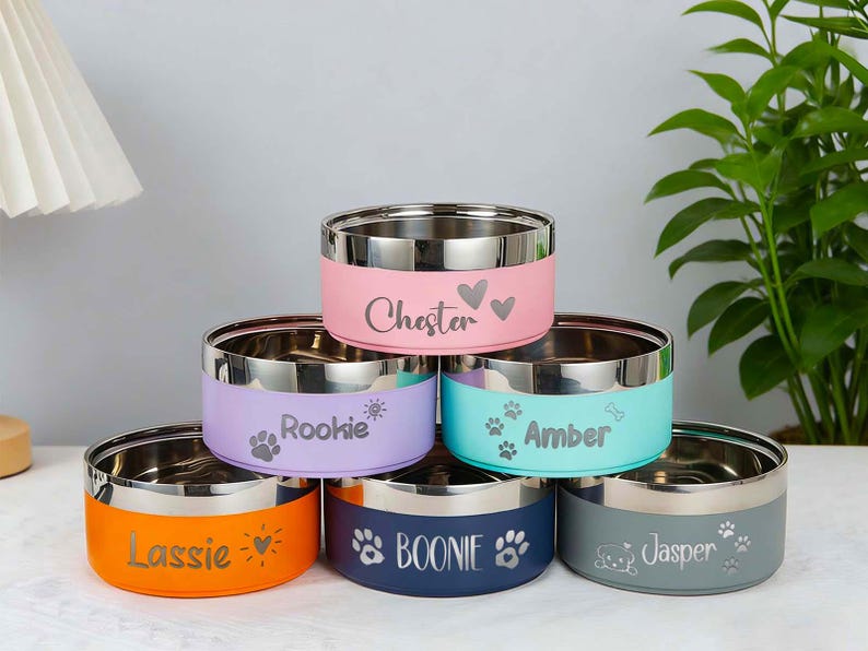 May include: A stack of six stainless steel pet bowls with colored exteriors. Each bowl has a different color: orange, navy, gray, pink, purple, and teal. Each bowl is personalized with a pet's name and paw print or heart design.