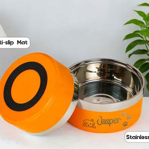 May include: An orange and stainless steel pet food bowl with the name "Jasper" and paw prints. The bowl has a black anti-slip mat on the lid. The words "Anti-slip Mat" and "Stainless Steel" are also visible.
