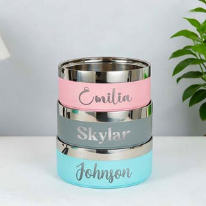 May include: Stack of three stainless steel food containers in pastel colors: pink, gray, and teal. Each container has a silver rim and a name engraved on the side: Emilia, Skylar, and Johnson. The containers are stacked on top of each other.