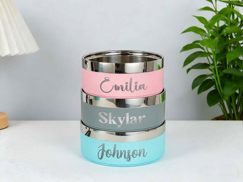May include: Stack of three stainless steel food containers in pastel colors: pink, gray, and teal. Each container has a silver rim and a name engraved on the side: Emilia, Skylar, and Johnson. The containers are stacked on top of each other.