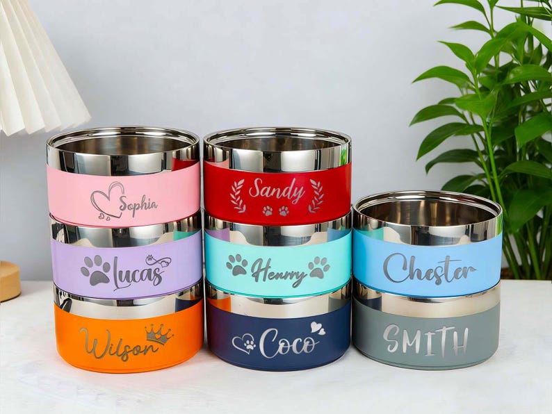 May include: A stack of six stainless steel pet bowls with colored bands. Each bowl has a different name engraved on it: Sophia, Lucas, Wilson, Sandy, Henry, Chester, and Smith. The bowls are in various colors, including pink, purple, orange, red, teal, blue, and gray.