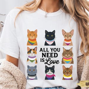 Puede incluir: Camiseta blanca con un diseño de nueve gatos de dibujos animados, cada uno con un pelaje diferente y una bufanda colorida. El texto "ALL YOU NEED IS Love" está impreso en negro. Cuello redondo.
