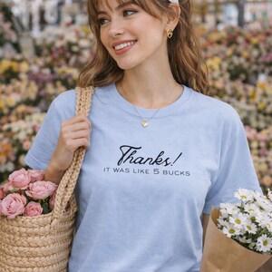 May include: A light blue t-shirt with the text "Thanks! It was like 5 bucks." A woman is holding a bouquet of white daisies and a woven tote bag filled with pink roses. She is wearing a white headband and gold earrings.