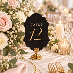 May include: A black, scalloped-edge table number sign featuring the gold text "TABLE 12" in a serif font. The sign is held upright by a small, circular, polished gold metal stand, creating a formal display piece for event seating arrangements.