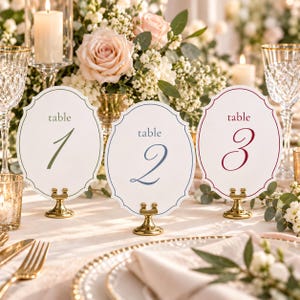 May include: Three oval table number cards with scalloped edges, each held by a small gold metal stand. The cards are labelled table 1 in olive green, table 2 in dusty blue, and table 3 in deep red, featuring elegant script numerals.
