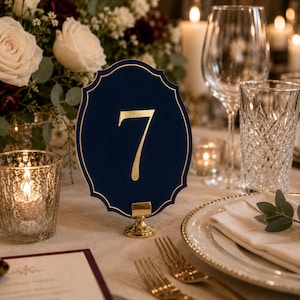 May include: A navy blue scalloped table number card featuring the gold foil number 7 in the center. The card is held upright by a small, round, polished gold metal stand, creating a formal display piece for a table setting.