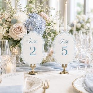 May include: Two scalloped-edge table number cards labeled Table 2 and Table 5, printed in blue on textured white cardstock. Each card is held upright by a small gold-toned metal stand, positioned on a table with floral arrangements.