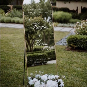 May include: A large, arched mirror with a gold frame serves as a wedding sign. The text "WELCOME TO THE WEDDING OF Kayla & Kevin" is displayed. The sign is decorated with white flowers and candles.