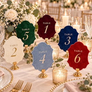 May include: A set of six scalloped velvet table number signs in cream, dark green, burgundy, dusty blue, navy blue, and red. Each sign features gold foil text reading TABLE followed by the corresponding number, mounted on a gold metal stand.