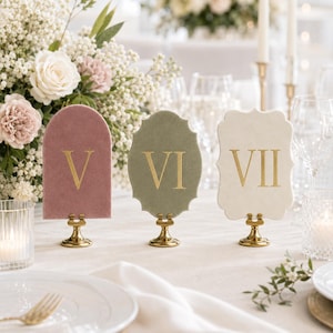 May include: Three velvet table number signs with gold Roman numerals V, VI, and VII. The signs are mounted on small gold metal stands. The first sign is arched and dusty rose, the second is scalloped and sage green, and the third is cream.