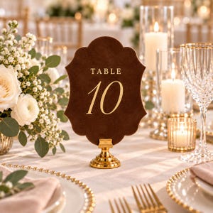 May include: A dark brown, scalloped-edge table number sign featuring the gold text "TABLE 10" centered on the surface. The sign is held upright by a small, polished gold metal stand with a circular base, positioned on a formal dining table.