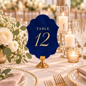 May include: A scalloped navy blue velvet table number sign featuring the gold text TABLE 12, mounted on a small gold pedestal stand. The sign has a smooth, soft-touch texture and is positioned upright to display the number clearly.