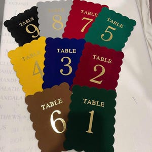 May include: A collection of nine rectangular table number cards with scalloped edges in various colors including black, gray, red, green, yellow, blue, and brown. Each card features the word TABLE and a large numeral in gold foil lettering.