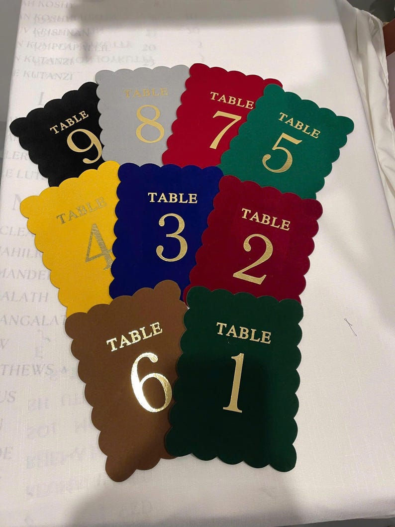 May include: A collection of nine rectangular table number cards with scalloped edges in various colors including black, gray, red, green, yellow, blue, and brown. Each card features the word TABLE and a large numeral in gold foil lettering.