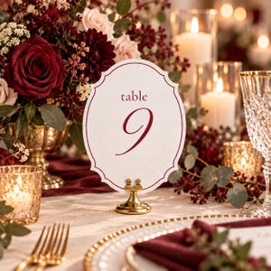 May include: A white scalloped table number card with a dark red border and the text table 9 in dark red script. The card is held upright by a small gold metal stand, positioned on a table with a floral arrangement and lit candles.