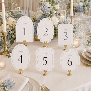 May include: A set of six white table number cards labelled one through six, each featuring a unique die-cut shape and black serif typography. The cards are displayed on small gold metal stands, arranged on a table with floral centrepieces.