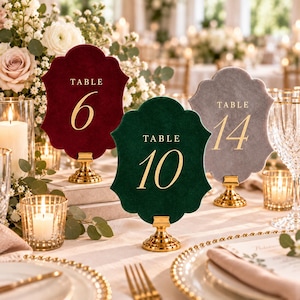 May include: Three scalloped-edge table number signs in deep red, dark green, and grey velvet. Each sign features gold-colored text reading TABLE followed by the numbers 6, 10, or 14, and is mounted on a small, circular gold-colored metal stand.