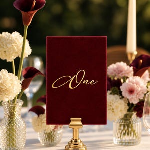 May include: A rectangular deep red velvet table number sign featuring the word One written in elegant gold script lettering. The sign is held upright by a polished gold metal stand with a tiered base, positioned on a white tablecloth.