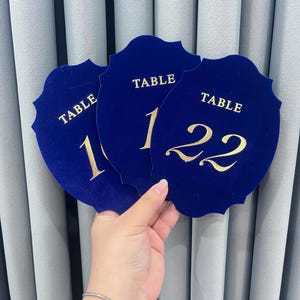 May include: Three shield-shaped table number signs made of deep blue velvet material. Each sign features the word TABLE in gold lettering above the numbers 10, 11, and 22 respectively. The signs have a scalloped edge design and a smooth texture.