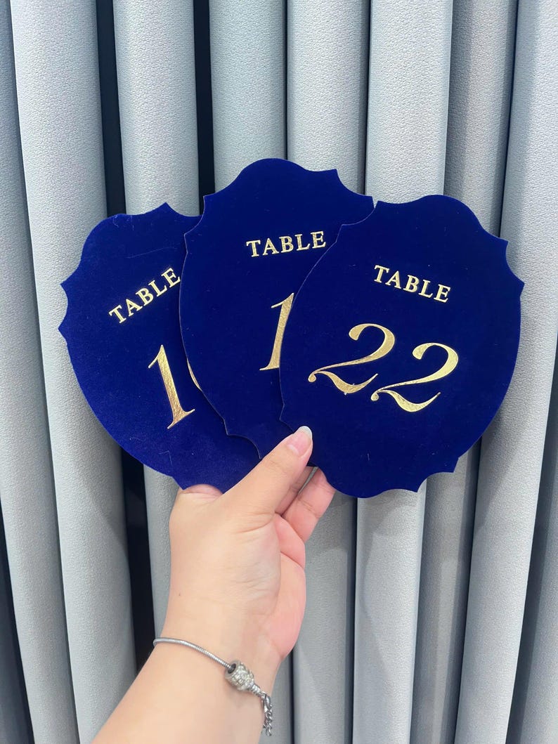 May include: Three shield-shaped table number signs made of deep blue velvet material. Each sign features the word TABLE in gold lettering above the numbers 10, 11, and 22 respectively. The signs have a scalloped edge design and a smooth texture.
