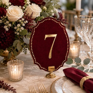 May include: A deep red velvet table number sign featuring the gold foil number 7 in the center, surrounded by a border of small gold stars. The sign has a scalloped edge and is supported by a small, round, polished gold metal stand.
