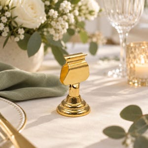 May include: A gold-colored metal place card holder with a circular base and a spring-loaded clip top. The stand features a polished, reflective finish and a vertical stem connecting the base to the clip, designed to hold small cards upright.