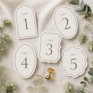 May include: Five off-white table number cards with sage green borders and text. The cards feature various shapes including an arch, oval, scalloped, and decorative frame. Each card displays the word TABLE and a large numeral from one to five.