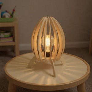 May include: A wooden table lamp with a unique design. The lamp features a light bulb encased in a light-colored, wooden, egg-shaped frame. The lamp is on a round table with three legs.
