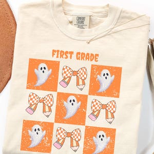 Puede incluir: Una camiseta de color crema con una etiqueta Comfort Colors en el cuello. El frente presenta el texto naranja FIRST GRADE sobre una cuadrícula de nueve cuadrados que contienen ilustraciones alternas de fantasmas blancos y lazos naranjas.