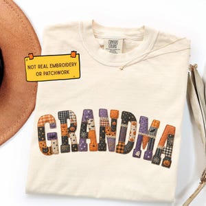 Puede incluir: Una camiseta color crema con la palabra GRANDMA en un estampado estilo patchwork con motivos de Halloween como calabazas, murciélagos y telarañas. Una nota amarilla en la camiseta dice: NOT REAL EMBROIDERY OR PATCHWORK.