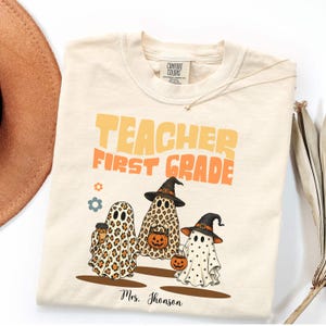 Puede incluir: Una camiseta color crema con el texto "TEACHER FIRST GRADE" en amarillo y naranja. Debajo, tres fantasmas ilustrados: uno con estampado de leopardo, otro con estampado de leopardo y sombrero de bruja, y otro con lunares.
