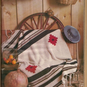 May include: A crocheted throw blanket with a striped pattern and red flower accents. The blanket is draped over a wooden wagon wheel and a rustic wooden chair.