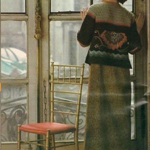May include: A gold bamboo chair with a red seat stands near a window. A person wearing a green, black, and orange crochet top and a long green skirt is looking out the window. The scene is indoors.